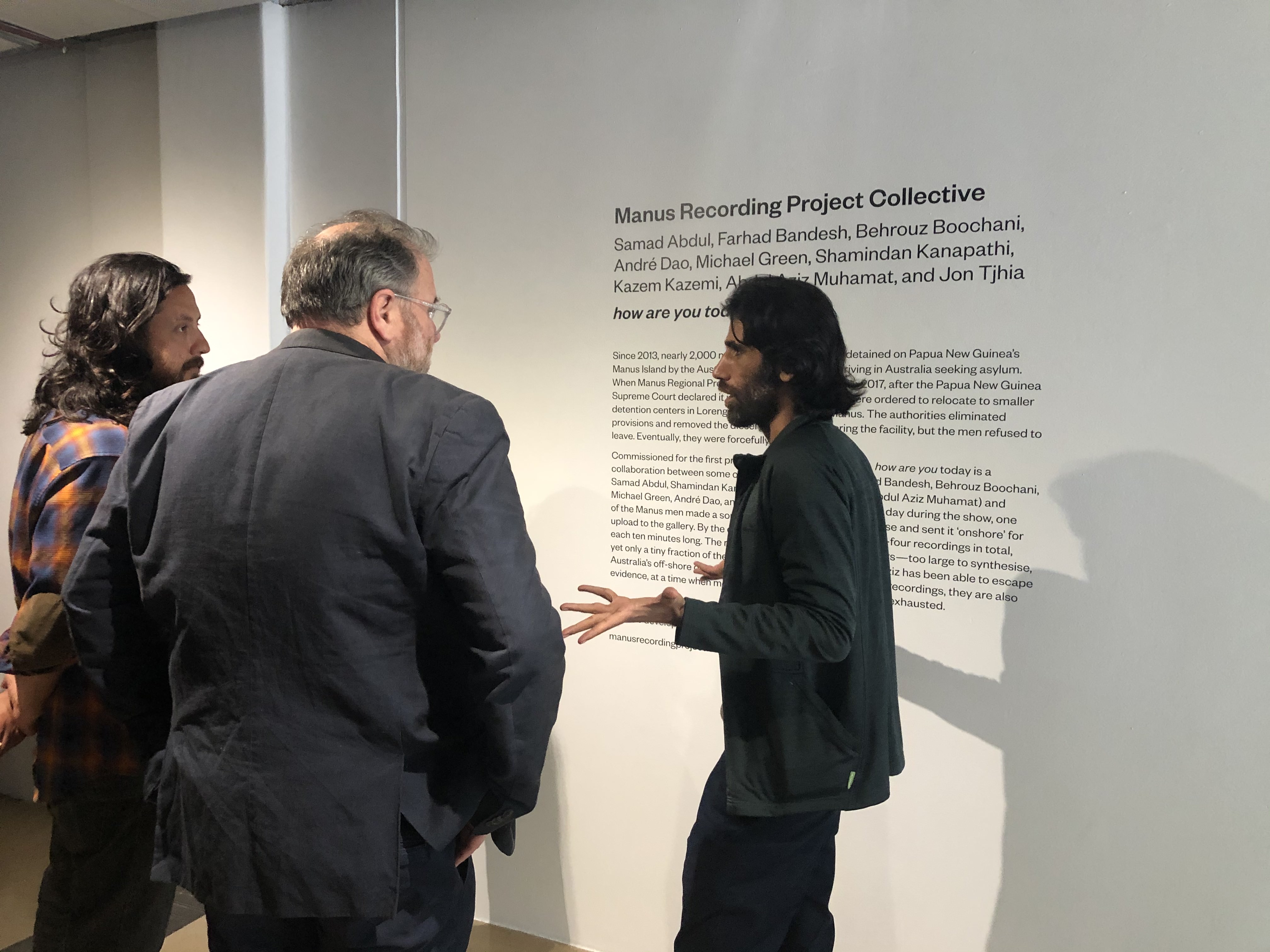 <p>Behrouz Boochani (right) with artist Bryan Philips and curator Robert  Leonard, next to the work <em>how are you today</em> by Manus Recording Project Collective, City Gallery Wellington, November 17, 2019.</p>