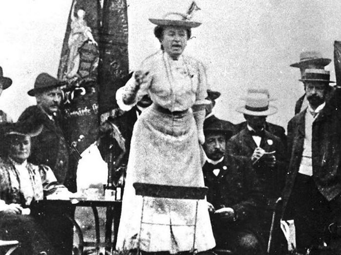 <p>A photo of Rosa Luxemburg speaking outside a socialist congress in 1907.<br>She is wearing a long dress, a wide-brimmed hat and is gesturing passionately. </p>